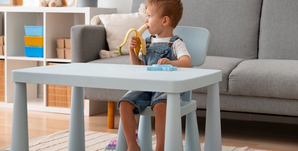 Un enfant mange tout seul une banane sur une petite table